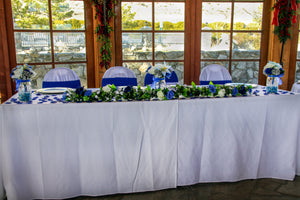 White tablecloths on a wedding head table with colorful flowers and chair sashes in front of a window