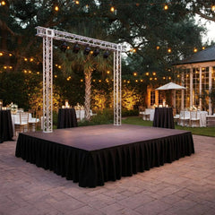 Outdoor event setup with a stage that has a black stage skirt and a truss on top. In the yard there are also dining tables, and string lights under trees.l