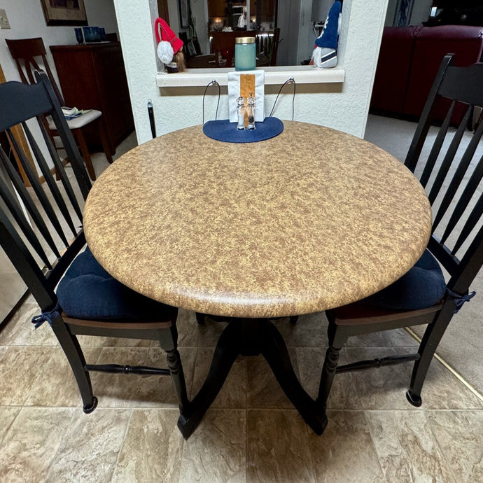 A round vinyl fitted tablecloth with a beige color pattern, placed on a table, with chairs around it, likely in a home setting.
