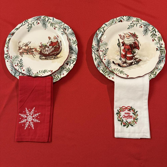 Embroidered Christmas napkins and holiday plates over a Christmas red tablecloth. One red napkin has a snowflake and the white napkins has Merry Christmas embroidered.