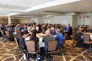Large group of people sitting at tables in a conference room setting. Tables have blank tablecloths