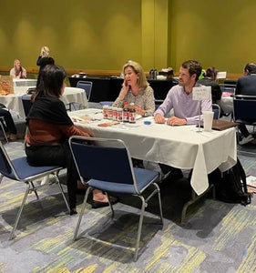 Attendees at a trade show sitting at vendors booth listening. Trade show table has a blank tablecloth