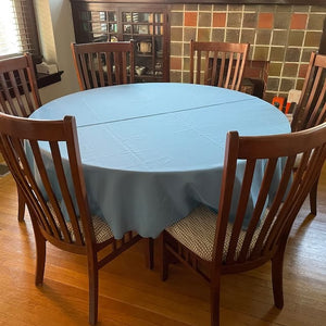 Round Table dressed with a light blue tablecloth in a home setting with 6 chairs in front of a window