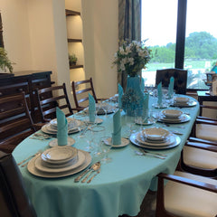 Turquoise oval tablecloth on a formal table setting with elegant china, silverware and glasses. The napkin fold is rolled and on top of dinner plates. There is also a ceramic vase and a beautiful white floral arrangement in the center.