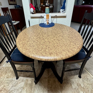 A round vinyl fitted tablecloth with a beige color pattern, placed on a table, with chairs around it, likely in a home setting.