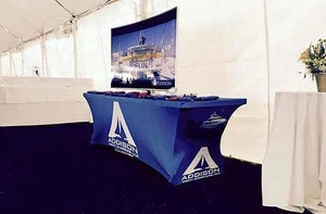Blue spandex table cover with white logo for Addison, set up with tv on top