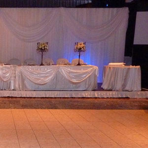 A white skirt on a wedding reception stage with a head table draped in fabric decor and flowers