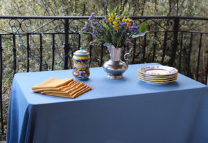 Light blue tablecloth set up outdoors with gold napkins, plates, and lavender flowers