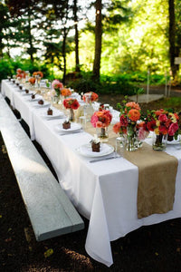 long Table with a white tablecloth and a natural colored runner at an outdoor reception with flowers