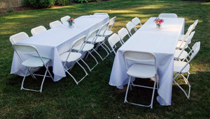 White rectangular table linens on event tables with foldout chairs in a backyard set up with flowers